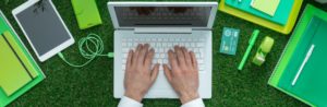 hands at a laptop sitting on top of a bed of grass with green office supplies surrounding it