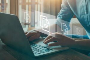 Icons of digital files hover above a close up of a young professional working on a laptop to signify document management