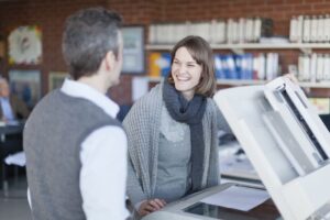 A businesswoman and businessman stand in front of an office multifunction printer making a copy.