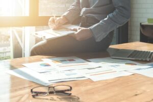 A closeup image of a man sitting at a desk with dozens of colorful printed sheets of paper sprawled around as he notates one on a clipboard signifying the importance of Managed Print Services.