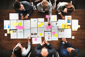 A top view of a diverse group of professionals sits around a table with colorful outputs spread neatly across the table signifying the importance of a small business copier.