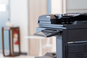The image shows a modern color copier in an office setting. The copier is prominent in the foreground, with a sleek design featuring a digital control panel illuminated with colorful buttons. The machine is equipped with multiple paper trays and a document feeder on top, indicating its multifunctional capabilities such as printing, scanning, and copying. The background is softly focused, featuring a neutral office environment with light-colored walls and minimalistic furniture. This color copier is likely a central piece of office equipment, used for creating vibrant, high-quality copies of documents and presentations.