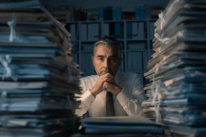 The image captures a man in professional attire, with a white shirt and tie, sitting at a desk and gazing thoughtfully through stacks of documents on either side of him. His chin rests on his clasped hands, and he seems to be surrounded by an overwhelming amount of paperwork. Behind him, shelves filled with binders suggest a storeroom or an office dedicated to keeping records. The man's expression conveys concern or contemplation, possibly considering strategies for document management to streamline the organization and handling of these numerous papers. The setting implies a need for an effective document management system to replace the physical clutter with a digital solution, improving accessibility and efficiency.