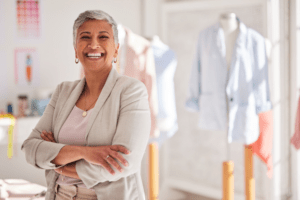 A cheerful, mature woman with short gray hair, wearing a blazer and standing confidently in a bright clothing boutique. Her crossed arms and joyful expression suggest pride and success. In the softly focused background, various garments are displayed, hinting at a creative small business environment. Although not visible in the image, a small business copier could be a useful addition to such a setting for handling paperwork and designs.