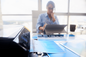 A businesswoman is focused on her laptop in a bright office, with a laser inkjet printer in the foreground and architectural blueprints spread out on the table.