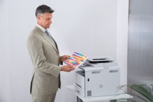 A man in a suit examining a color printout next to an office copier, showcasing the copier's capability to produce high-quality color documents for professional use.