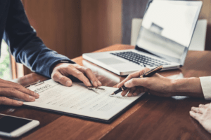 Close-up of two business professionals reviewing and signing a contract on a clipboard, with a laptop and smartphone on the table. Copier rental agreement in progress.