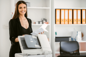 Smiling businesswoman using a printer in an office setting, holding printed documents. Managed print services ensuring efficient document handling.