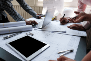 Group of professionals working on architectural blueprints at a desk with a laptop, tablet, and rolled-up plans. Wide format printing in use for detailed project designs.