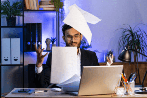 A frustrated office worker tosses papers into the air while sitting at a desk with a laptop, highlighting the need for effective document management. The background includes shelves with books and plants in a modern office setting.