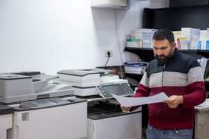 A small business owner reviewing a printed document in front of s small business copier. The office space is equipped with modern printing machines, shelves filled with invoices, and office supplies, reflecting a typical printing and copying setup for small businesses.