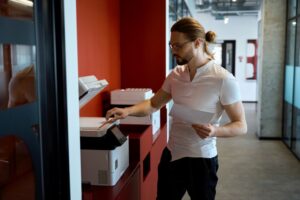 A professional in a modern office setting using a multifunctional color copier to scan or print a document. The copier is positioned against a red wall, and the individual holds a sheet of paper while operating the machine, highlighting the functionality and efficiency of color copiers in a workplace environment.