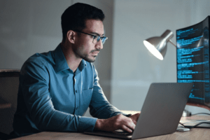 A focused IT professional working on a laptop in a dimly lit office, with a curved monitor displaying lines of code in the background. The image highlights the expertise and dedication provided by Managed IT Services in maintaining secure and efficient technology solutions for businesses.