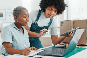 Two women working in an office, using a laptop and tablet to manage shipping tasks, with a postage meter and packing materials in the background.