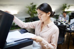 A young professional stands in front of a color office copier reviewing outputs while colleagues work behind her.