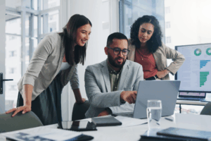 A group of young professionals stand around a laptop with a screen of charts and graphs in the background signifying Business IT Management