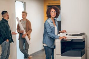 A woman stands in front of a business copier pushing a button on the interface while coworkers chat in the background.
